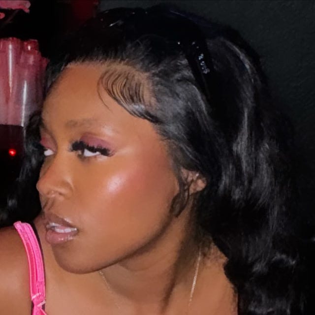 A close-up of a woman with soft, glowy makeup, long curled black hair, and styled baby hairs. She wears a pink spaghetti strap top and has a confident, slightly side-eyed expression. The background is dimly lit with a red glow, suggesting a night-out setting.