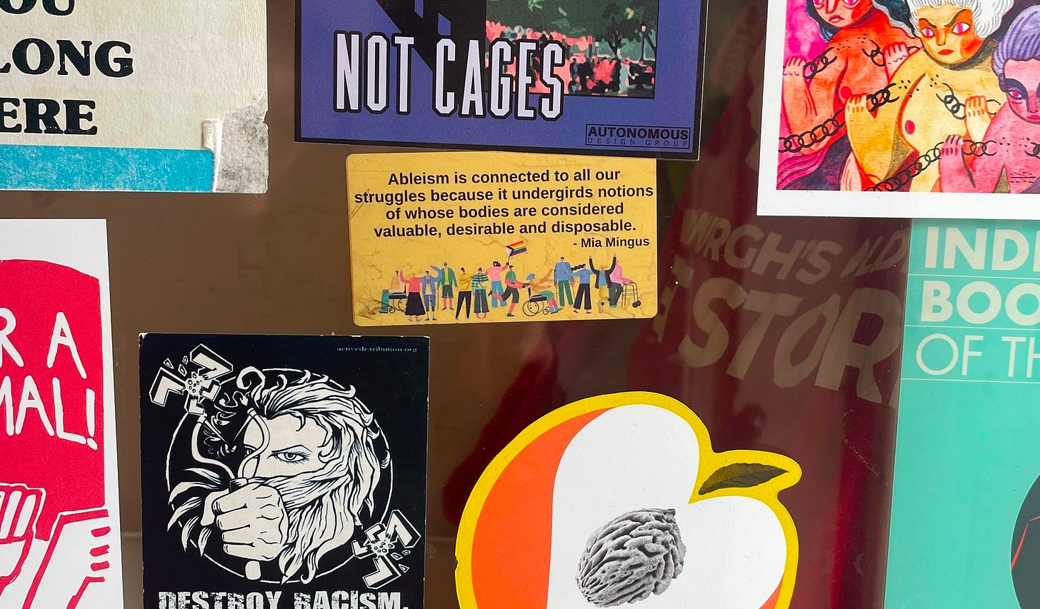 A shop window covered in stickers. The one in the middle says: Ableism is connected to all our struggles because it undergirds notions of whose bodies are considered valuable, desirable, and disposable." - Mia Mingus. The other stickers are cut off. 