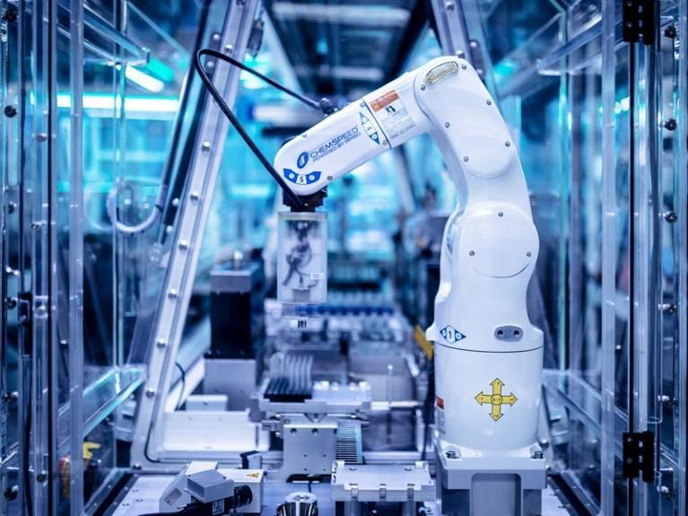 A robotic arm inside an enclosed, automated laboratory handling sample racks — the kind of remotely operated 'cloud lab' system that executes experiments without a human ever entering the room. A robotic arm inside an enclosed, automated laboratory handling sample racks — the kind of remotely operated 'cloud lab' system that executes experiments without a human ever entering the room.