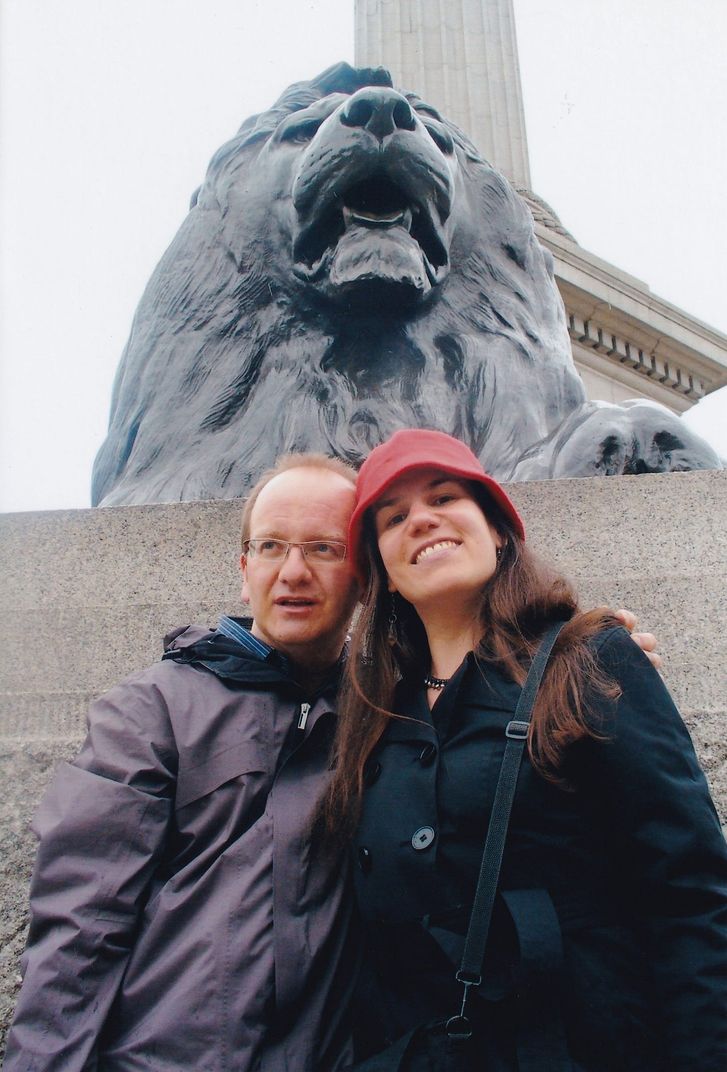 A man and a woman stand together with one of the stone lions from Trafalgar Square behind them. She is smiling at the camera and he is smiling as he looks to the side. 