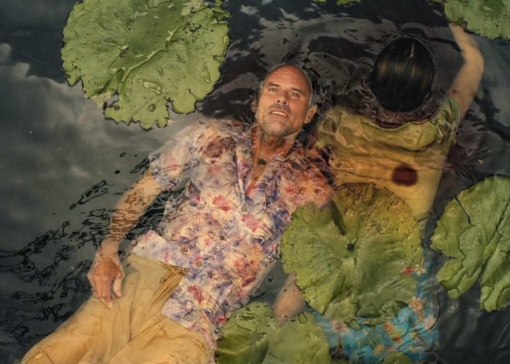 A photo of a dead couple lying amid water lilies in a dark pool of water. He is face up in a floral printed shirt, staring blankly into the sky, and she is face down.