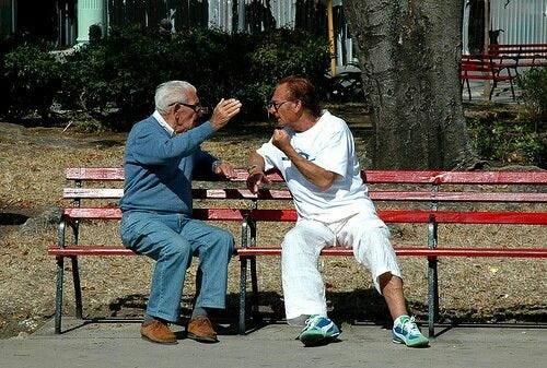This may contain: two older men sitting on red benches in front of a tree and one is holding his hand up to the other man's face