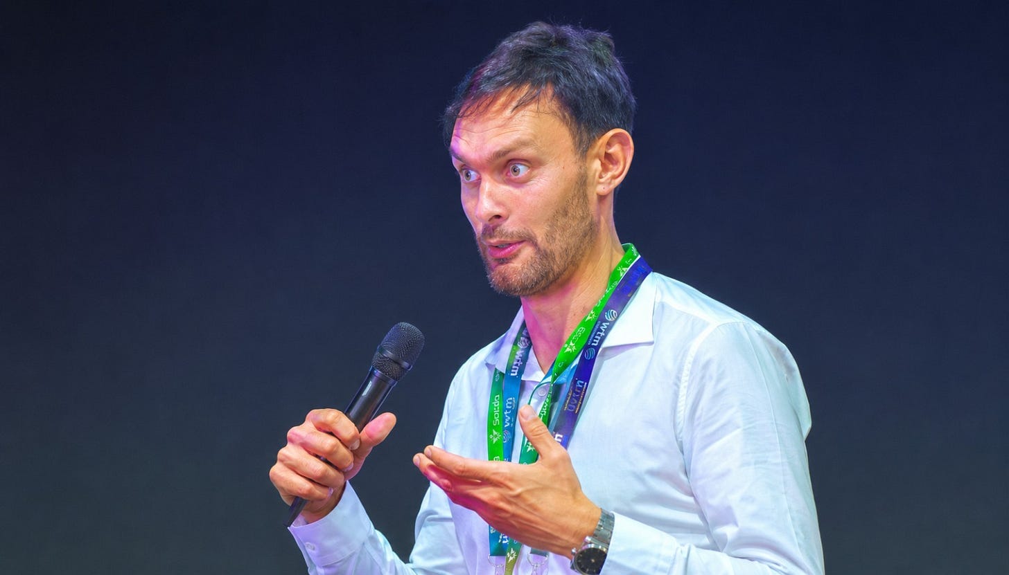 Harry Smith, a white man with brown hair and a brown beard, is wearing a white shirt and a green lanyard, speaking into a microphone held in his right hand while gesturing with his left hand.