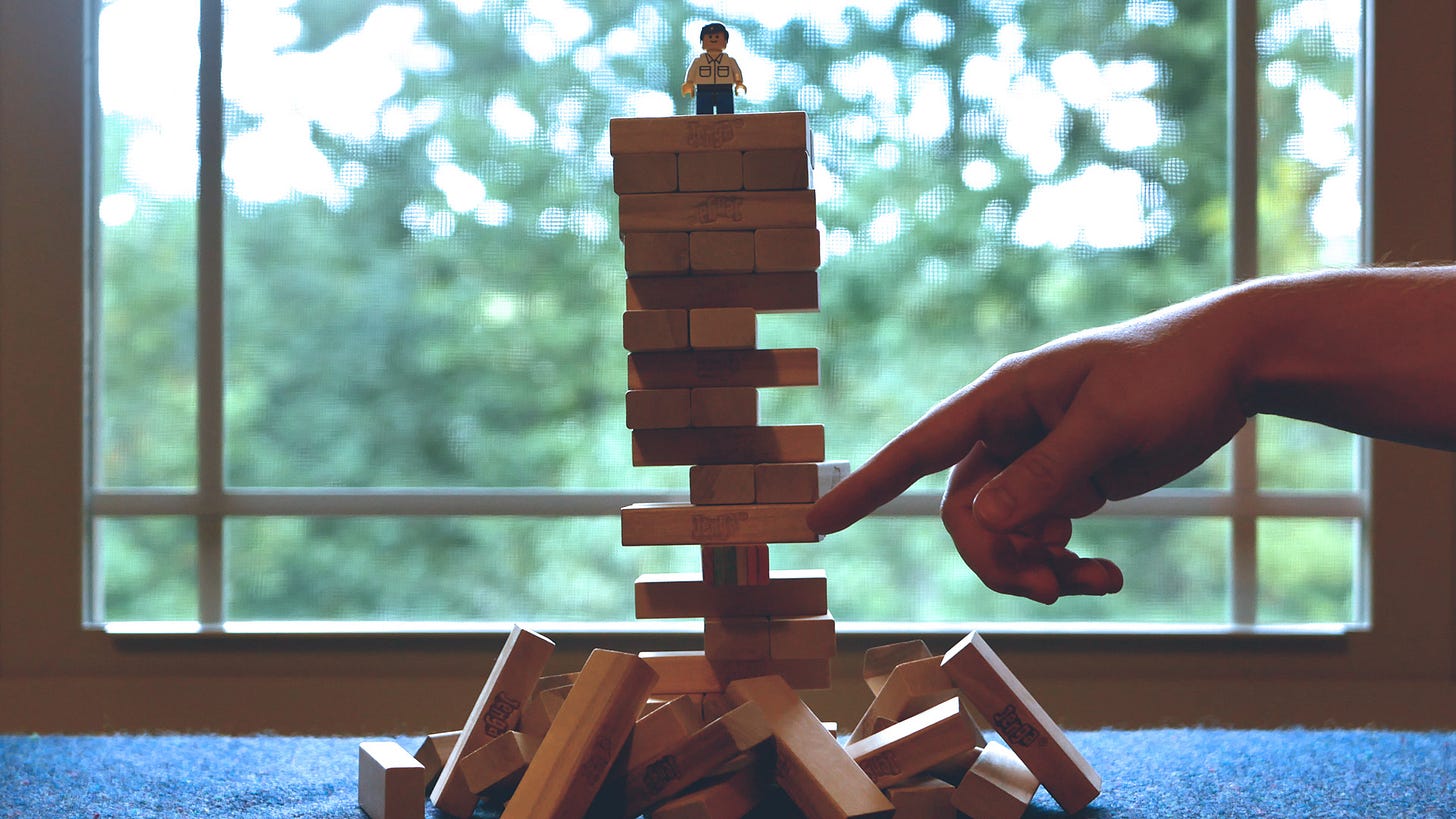 A male LEGO minifigure stands on top of a Jenga tower with many pieces in a pile around the base. It's in front of a window looking outside to a blurry view of trees. A hand reaches in from the right side of the image, loosely pointing at a piece with the colors of the rainbow on its side.