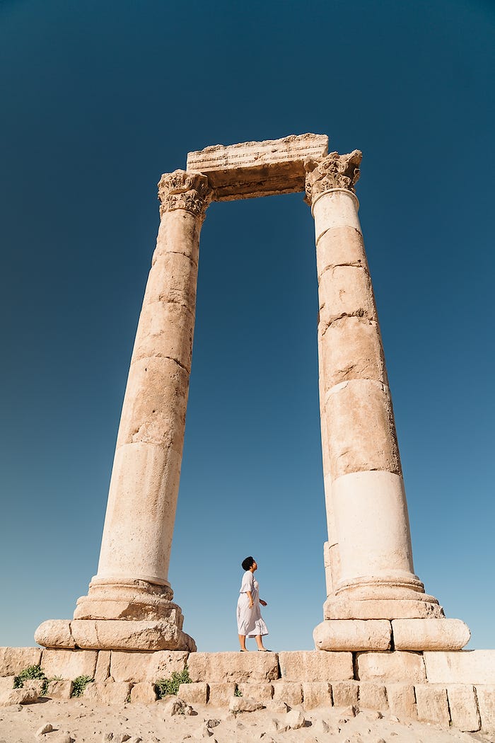 Anela stands in a midi dress in the sun underneath a stone archway at the Citadel in Amman Jordan, a site with Roman, Byzantine, and Umayyad ruins