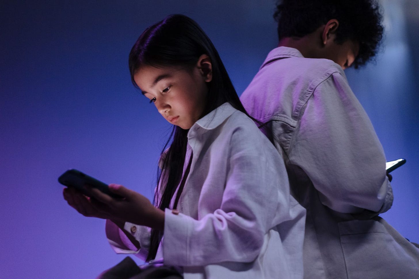 Two children sitting side by side, each absorbed in their own smartphone in a dimly lit room Two children sitting side by side, each absorbed in their own smartphone in a dimly lit room