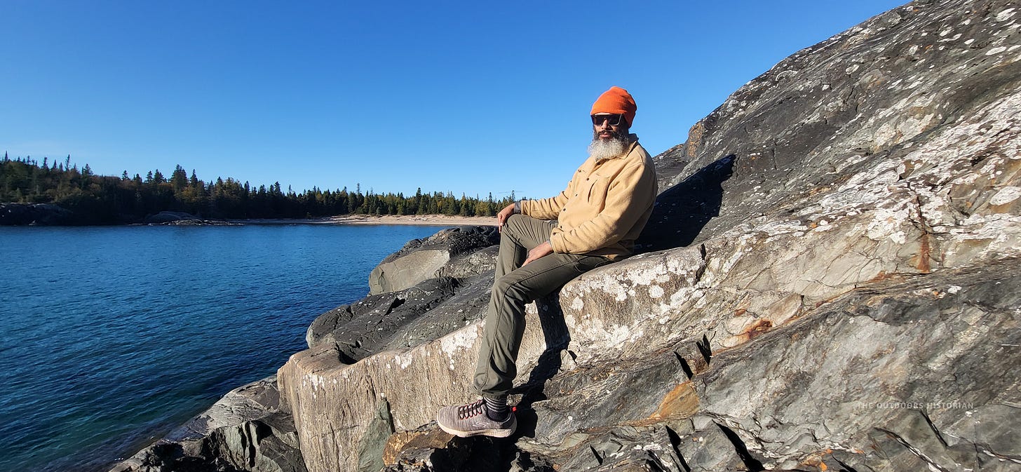Eugene sitting peacefully on a large boulder along the shoreline. He is wearing sunglasses, a bright orange toque, a beige jacket, and grey hiking pants, with a distant sandy beach and forest behind him.