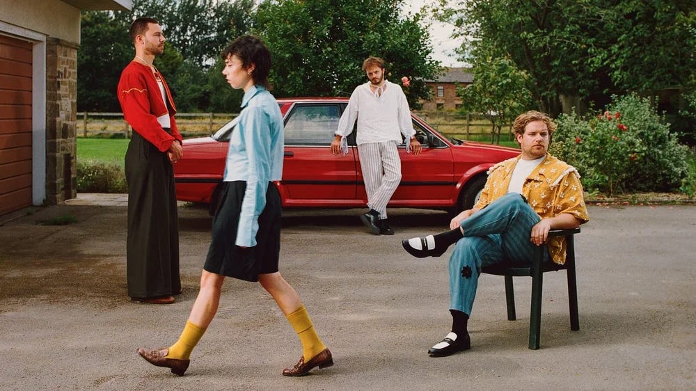An image from a Divorce photoshoot. Felix rests against a red car from the 1990s. Kaspar sits in a plastic chair and looks at the camera. Adam stands facing to the right, wearing a cropped red cardigan and wide-legged black trousers. Tiger walks from right to left, blurred by her movement.