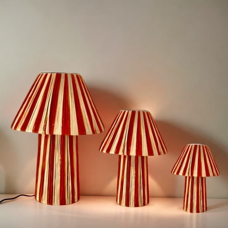 May include: Three red and cream striped mushroom lamps of varying sizes. The largest lamp is on the left, the medium-sized lamp is in the center, and the smallest lamp is on the right.  All three lamps have a similar design with a wide, pleated shade and a cylindrical base. The lamps are illuminated, casting a warm glow. These decorative table lamps are perfect for adding a touch of whimsy and style to any room.