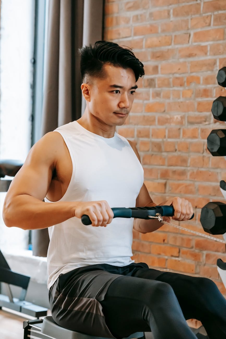 Man exercises on rowing machine in gym. Wearing white tank top and black pants. Brick wall and dumbbells in background. Focused expression.