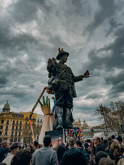 A gallery of Fallas monuments in Valencia, Spain — elaborately sculpted figures including a Charlie Chaplin figure holding butterflies, fantasy warriors, and colorful street sculptures, photographed against dramatic cloudy and blue skies in March 2026.
