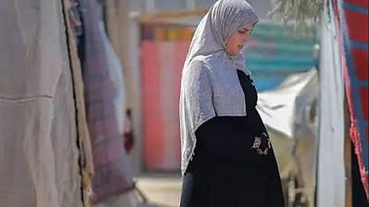 A side-profile view of a person wearing a light grey hijab and a long black dress or abaya, walking through a narrow outdoor passage lined with canvas tents or temporary shelters. The background is softly blurred, with muted colours and fabric structures suggesting a camp or informal settlement. The person is looking downward with a neutral, contemplative expression.