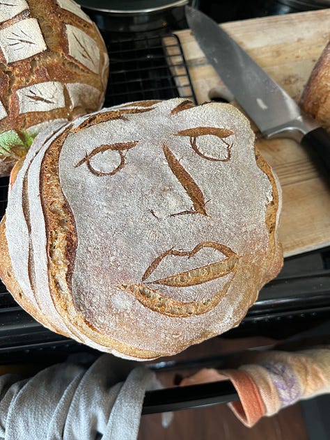 three round loaves of sourdough with different beautiful scoring techniques used. one is a lovely grid with wheat sheaves, one is a portrait of a face, another is an intricate swirl