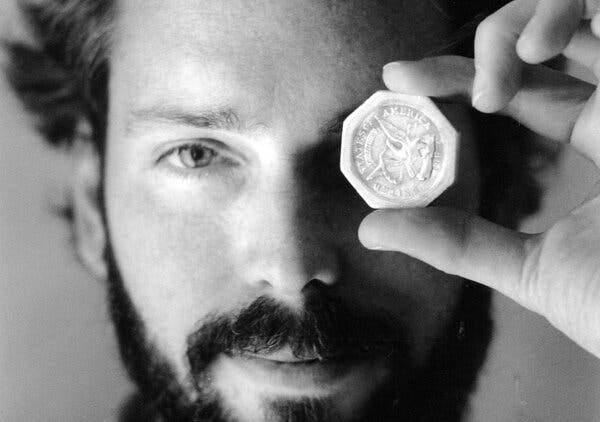 Un hombre con barba se acerca una moneda de oro al ojo.