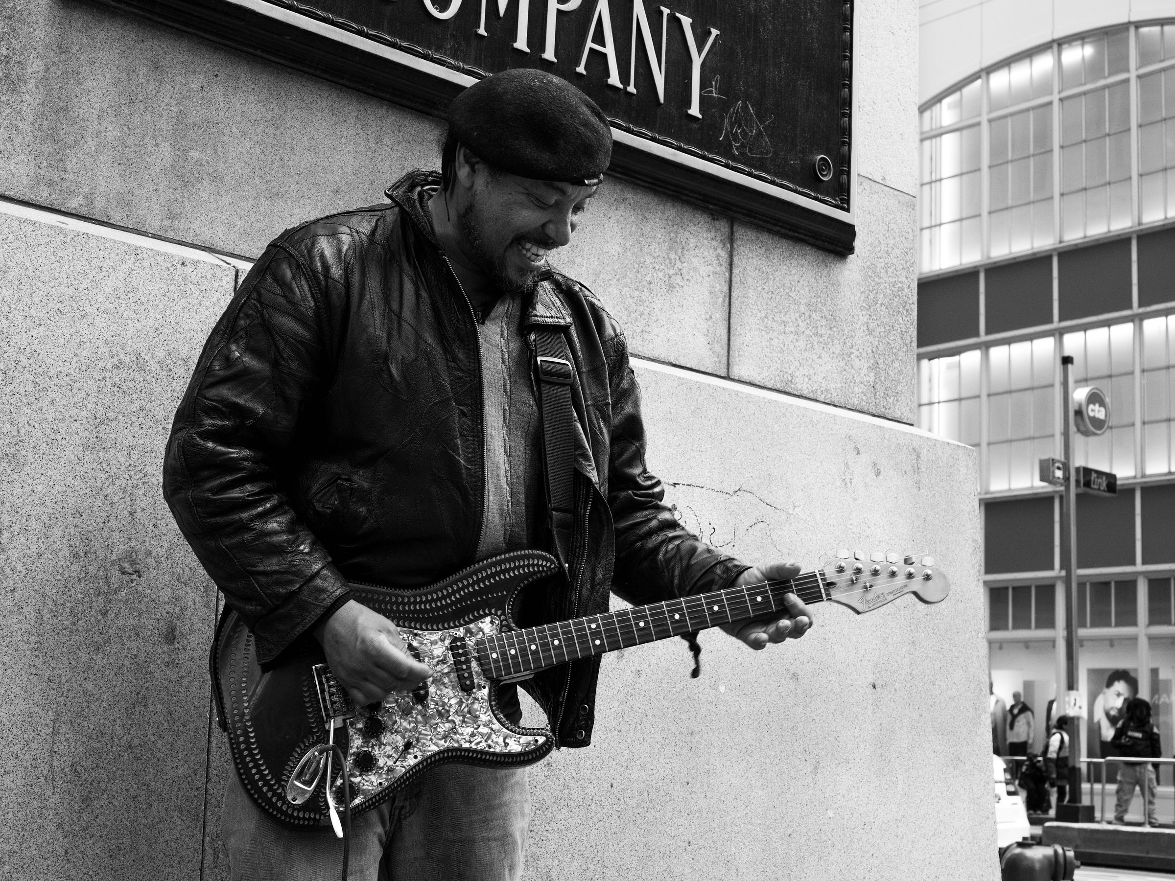 Black and white photograph of a street musician playing an electric guitar on a city sidewalk. He wears a leather jacket and a beret, smiling as he strums. Behind him, a CTA sign and modern glass buildings frame the urban backdrop, with pedestrians passing by. Black and white photograph of a street musician playing an electric guitar on a city sidewalk. He wears a leather jacket and a beret, smiling as he strums. Behind him, a CTA sign and modern glass buildings frame the urban backdrop, with pedestrians passing by.