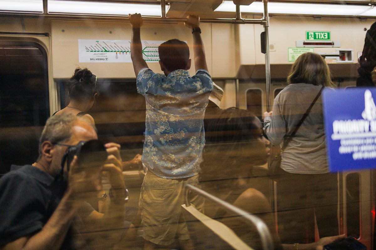 David Rosen Self Portrait Reflection and Interior view of a subway car showing a man in a Hawaiian shirt holding onto the overhead rail, surrounded by seated and standing passengers, with reflections in the glass. David Rosen Self Portrait Reflection and Interior view of a subway car showing a man in a Hawaiian shirt holding onto the overhead rail, surrounded by seated and standing passengers, with reflections in the glass.