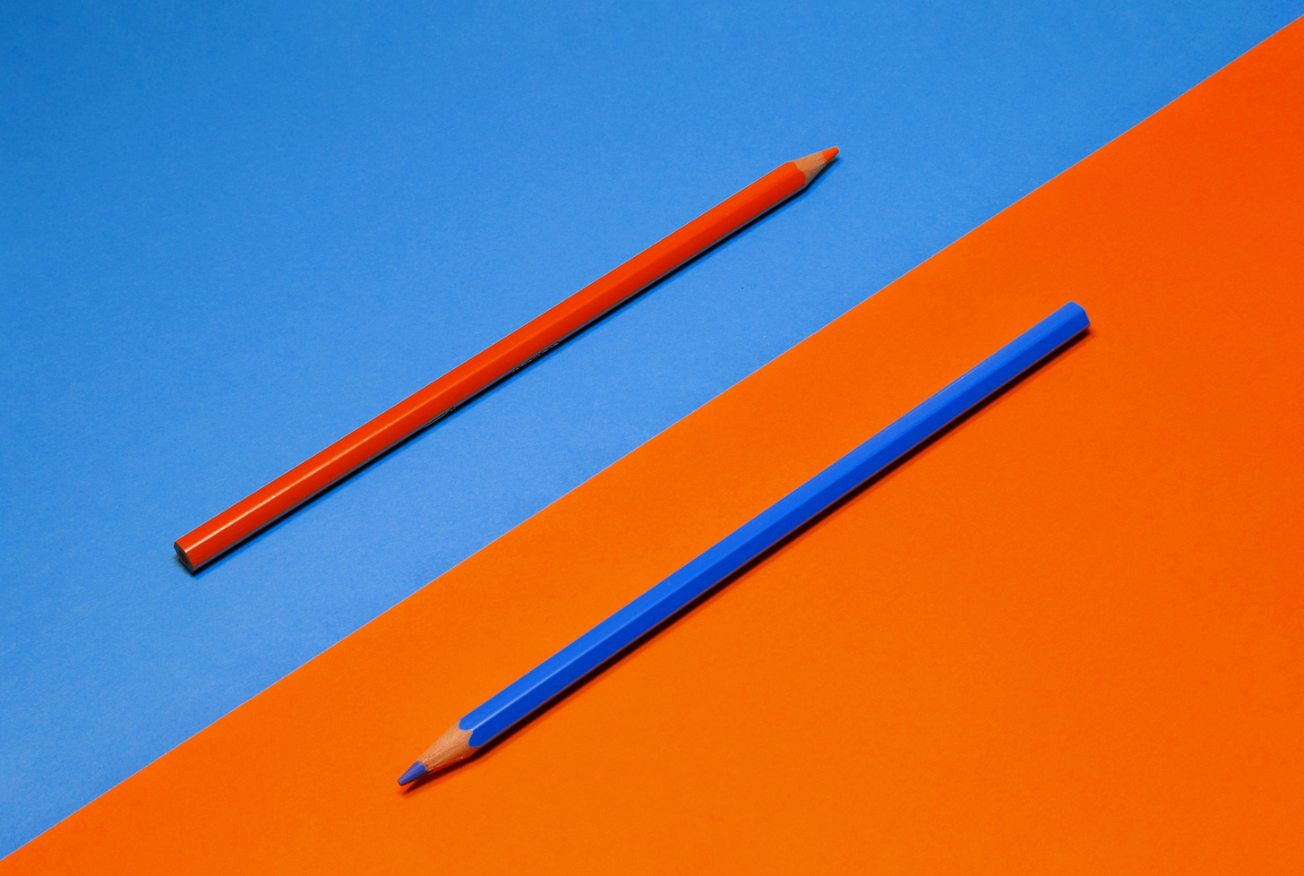 Two colored pencils—one orange on a blue background and one blue on an orange background—placed diagonally across contrasting colored paper.