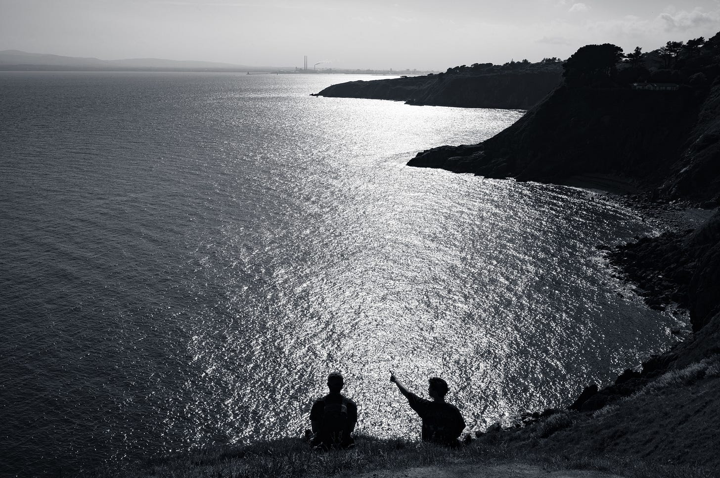 Two figures silhouetted on a clifftop looking out over a bay of glittering sunlit water, in black and white, a headland running into the distance. Two figures silhouetted on a clifftop looking out over a bay of glittering sunlit water, in black and white, a headland running into the distance.