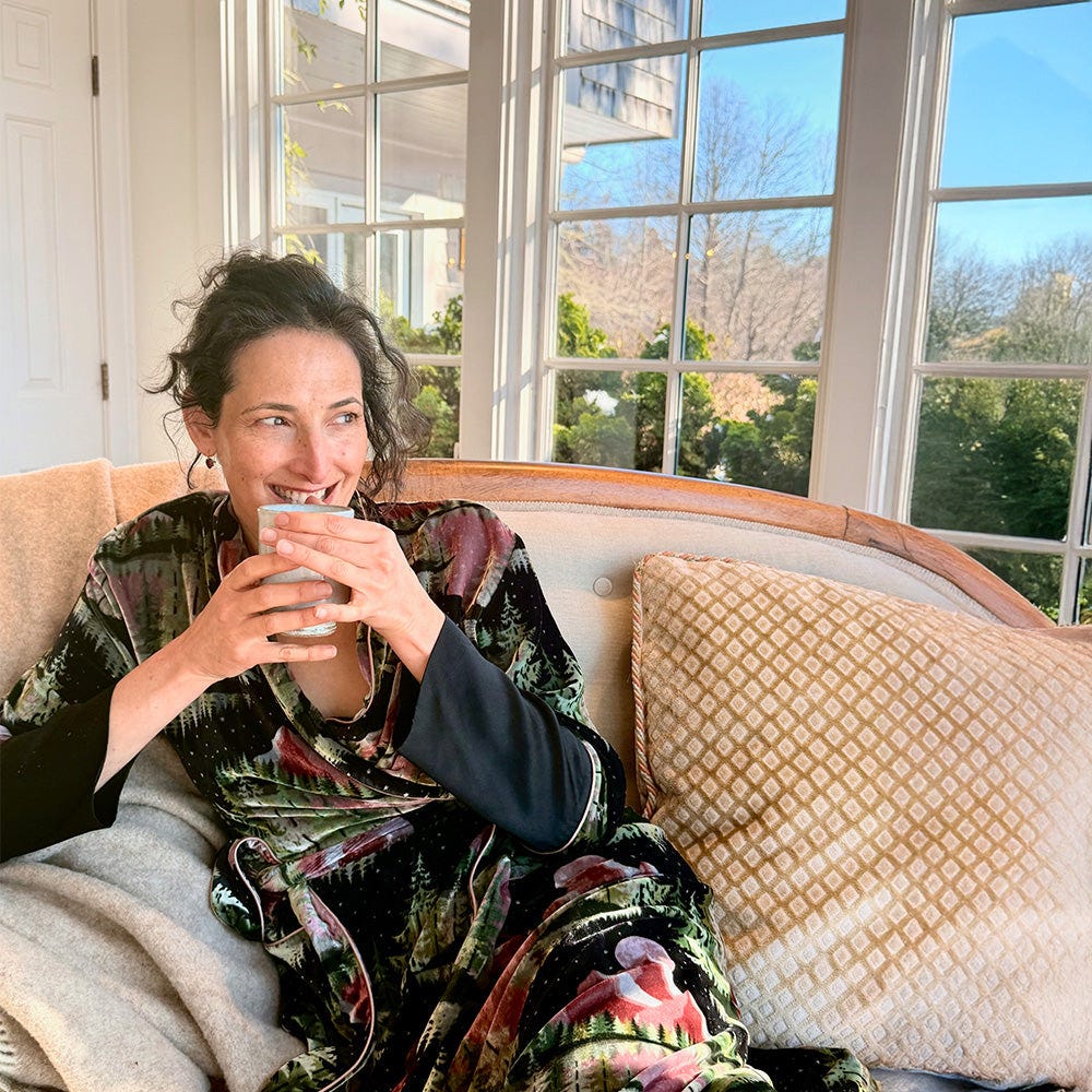 Dr. Catherine sitting on a couch holding a cup of tea, with large windows showing a scenic view outside. Dr. Catherine sitting on a couch holding a cup of tea, with large windows showing a scenic view outside.