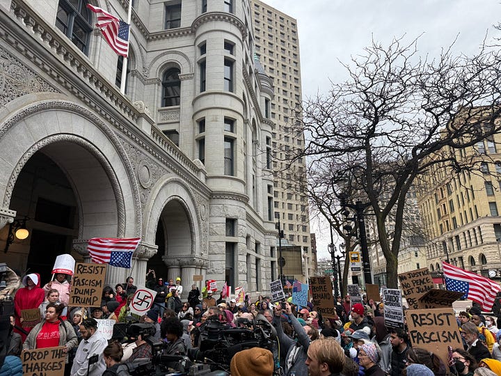 A crowd in front of a federal building with an arched entryway. THey are holding signs, including a State of LIberty made up as Rosie the Riveter saying "STOP THE ABUSE OF POWER" and quoting the Due Process clause of the Constitution the picture on the left and "Resist Fascism, Dump Trump," an upside-down US flag and "Drop The Charges" on the right.