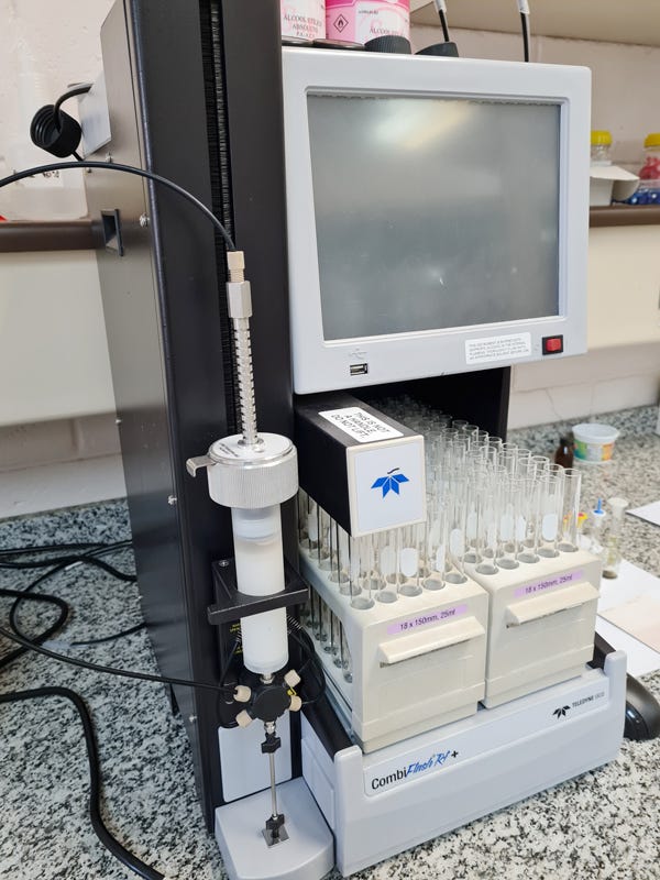 Equipamento de análise laboratorial automatizado com monitor branco de tela cinza acoplado na parte superior, base preta contendo compartimentos com tubos de ensaio transparentes organizados em suportes brancos, frasco de coleta de amostra branco acoplado ao lado esquerdo, caixa branca com logotipo azul posicionada entre os suportes de tubos, fios e cabos pretos conectados ao equipamento, superfície de trabalho em granito cinza ao fundo com prateleiras brancas contendo frascos e materiais diversos, e headphones pretos pendurados na lateral esquerda do equipamento. Equipamento de análise laboratorial automatizado com monitor branco de tela cinza acoplado na parte superior, base preta contendo compartimentos com tubos de ensaio transparentes organizados em suportes brancos, frasco de coleta de amostra branco acoplado ao lado esquerdo, caixa branca com logotipo azul posicionada entre os suportes de tubos, fios e cabos pretos conectados ao equipamento, superfície de trabalho em granito cinza ao fundo com prateleiras brancas contendo frascos e materiais diversos, e headphones pretos pendurados na lateral esquerda do equipamento.