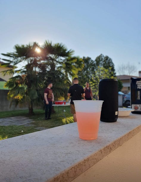 Image 1: A picturesque stone bridge crossing a Venetian canal at sunset, with people walking across and reflections of historic Italian architecture shimmering in the blue water. A small boat is docked on the left, and a terracotta building with white trim stands on the right. Photo taken in Venice, Italy. Image 2: Outdoor summer gathering in an Italian backyard, featuring a plastic cup with a pink cocktail in the foreground, palm trees, and a group of friends playing beer pong in the background. The sun is shining behind the trees, giving a relaxed Italian party vibe. Photo taken in Italy. Image 3: Classic Venetian canal scene with colorful historic buildings, a moored boat, and calm water reflecting the architecture. The canal stretches toward a distant clock tower under a bright blue sky. Photo taken in Venice, Italy. Image 4: Black-and-white photograph of Venetian canal houses and rippling water, highlighting the texture and timeless character of Italy’s iconic canal-side architecture. Taken in Venice, Italy. Image 5: Close-up of a gray cat perched on a stone ledge with lush Italian greenery behind, while another cat lounges on the pavement next to a blue car. The evening sky is partly cloudy, capturing a quiet residential moment in Italy.