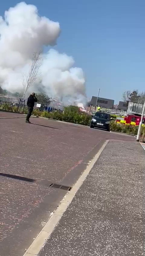 Large plume of smoke rising from a building fire; firefighters and a bystander are visible.