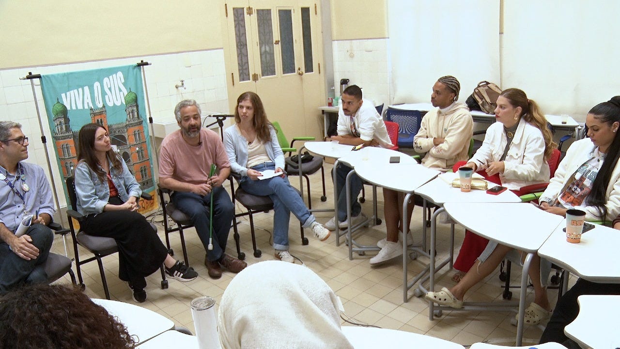 Em uma sala com paredes claras e piso bege, a foto registra parte de uma roda de conversa. José Carlos, Daniele Novaes, Manoel Negraes e Clara Vale estão sentados lado a lado em cadeiras pretas com rodinhas. Ao fundo, há um cartaz com a frase "Viva o SUS" e a imagem do Castelo Mourisco da Fiocruz. À direita da imagem, aparecem quatro influenciadores, duas mulheres e dois homens, sentados em cadeiras com mesas escolares brancas. No canto inferior direito, vê-se parcialmente a parte de trás da cabeça de duas pessoas (fim da descrição)