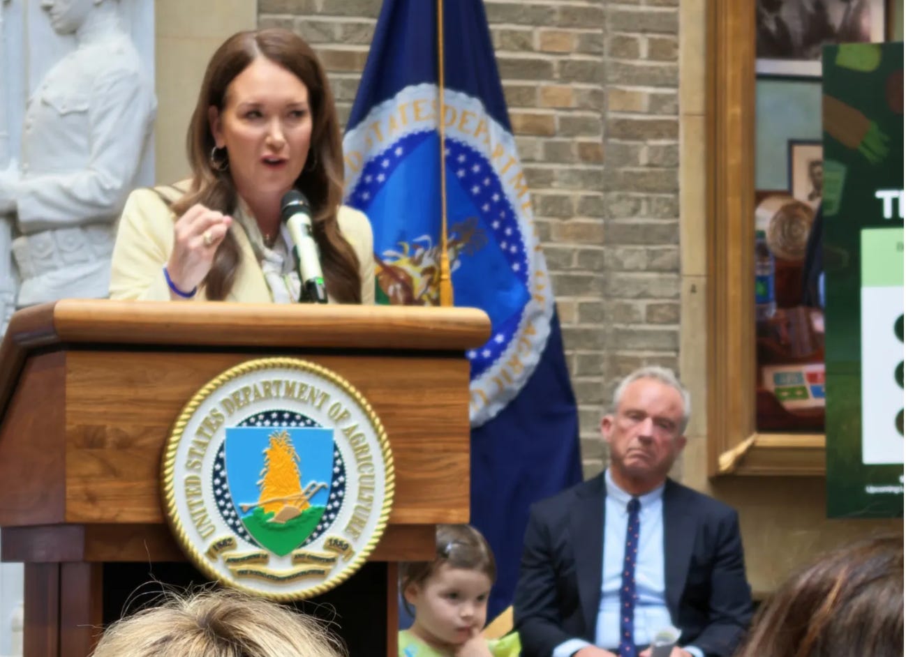 Brooke Rollins speaks at a USDA podium as Robert F. Kennedy Jr. sits behind her at the announcement