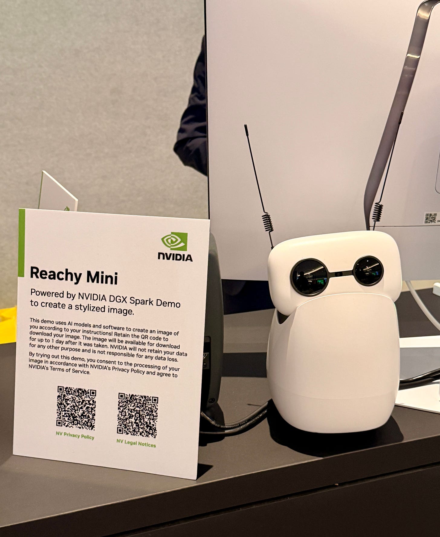 NVIDIA’s Reachy Mini robot is displayed on a counter at CES beside an informational sign describing the demo. The small white desktop robot has round black eyes and antenna-like sensors. NVIDIA’s Reachy Mini robot is displayed on a counter at CES beside an informational sign describing the demo. The small white desktop robot has round black eyes and antenna-like sensors.