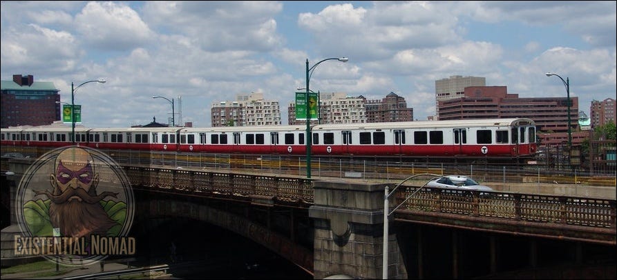 This is a photograph of a red and white subway train, part of the MBTA's Red Line, traveling across a metal bridge. The train is elevated, and below the bridge, there is a street. In the background, there is a cityscape with multiple buildings, some of which appear to be brick, and a partly cloudy, blue sky above.