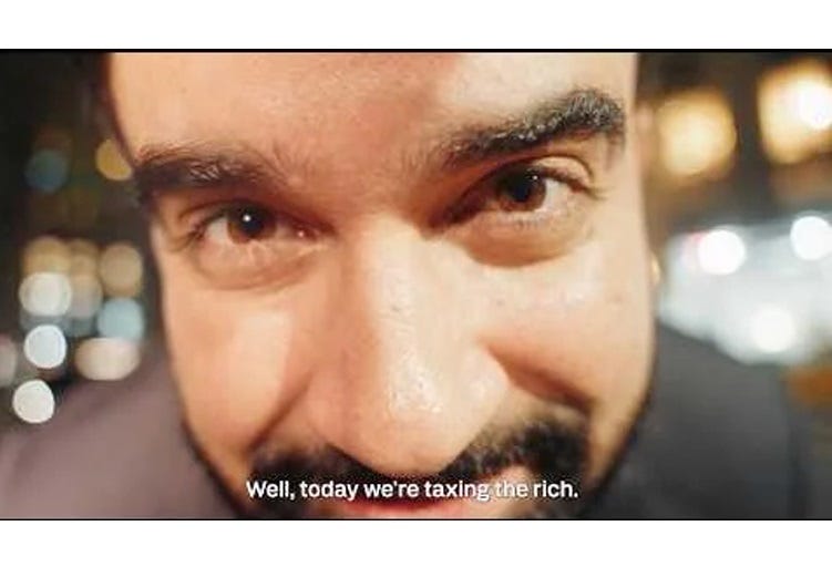 Video screenshot: A smiling Zohran Mamdani leans down to put his face extremely close to the camera lens, filling most of the frame. Caption: 'Well, today we're taxing the rich'