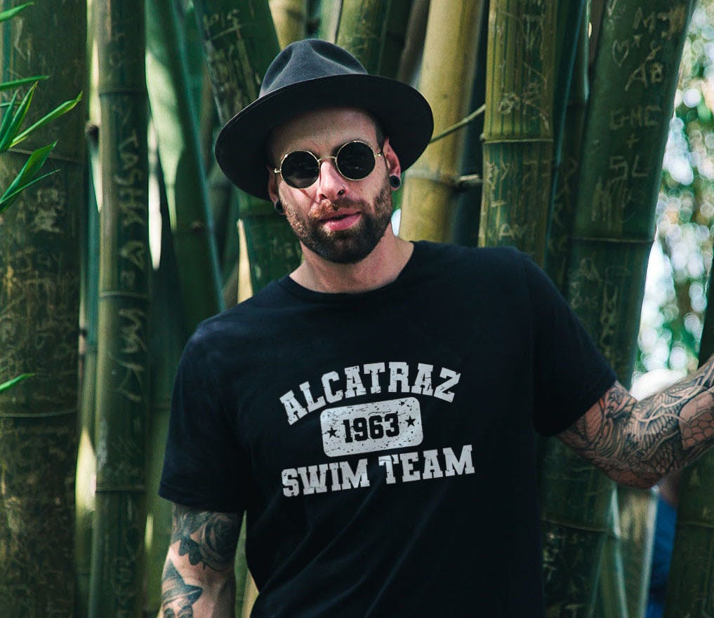 Male hipster with fedora wearing a black t-shirt with printing that reads Alcatraz Swim Team