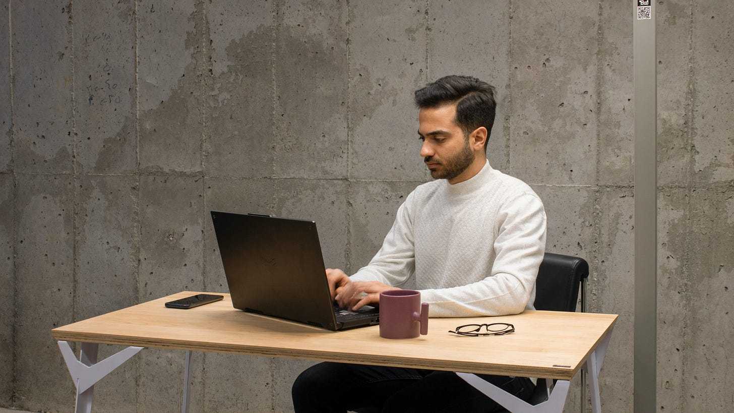 Focused individual working on a laptop at a minimalist desk with a concrete wall backdrop, accompanied by a mug and glasses, symbolizing concentration and productivity in a modern workspace.