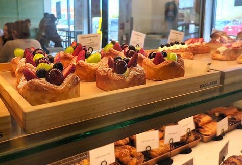 Tous les Jours display case items including fruit-loaded danishes, corn cheese donuts coated in granulated sugar, and a variety of croquettes including mango and chocolate