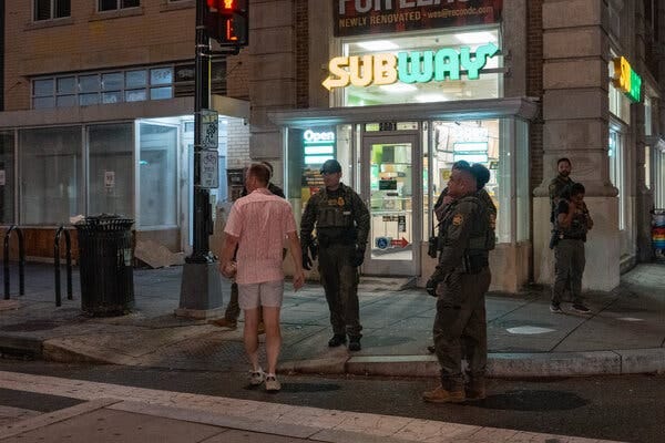 A man in a pink shirt holding something in his left hand and several people in law enforcement uniforms stand outside a Subway sandwich shop on a city street corner.