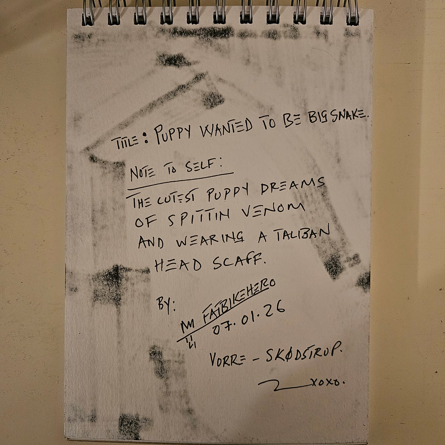 A photograph of a sketchbook page with handwritten notes in black ink. The text reads: "Title: PUPPY WANTED TO BE BIG SNAKE." Below, a "Note to self" states: "The cutest puppy dreams of spittin venom and wearing a Taliban head scaff [scarf]." The page is signed "By: M FatbikeHero," dated "07.01.26," and notes the location "Vorre - Skødstrup."