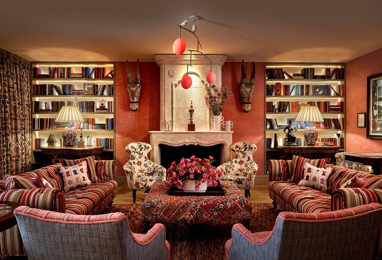 The Drawing Room at Warren Street Hotel with two sofas upholstered in a dark red striped fabric, a large rectangular footstool in a red embroidered fabric and two curvaceous statement armchairs in a cream, red and blue floral fabric. In the background is a stone fireplace with bookshelves on either side