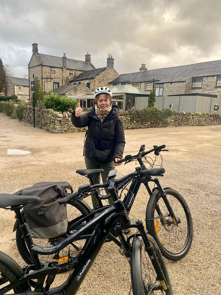 Two photos side by side. On the left, a woman stands beside an e-bike outside stone cottages in a Peak District village, smiling and giving a thumbs up. On the right, the same woman wears protective clothing and a helmet inside a workshop, standing next to machinery while making jewellery.