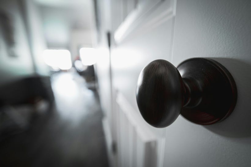 A close-up of a round brass doorknob in a white door, at an angle so the door is to the viewer's right. Beyond the door, the room is blurred, making it hard to tell what's happening: there's maybe a wood floor, some kind of table, a couple of wall lamps? The uncertainty communicates a certain ominousness. A close-up of a round brass doorknob in a white door, at an angle so the door is to the viewer's right. Beyond the door, the room is blurred, making it hard to tell what's happening: there's maybe a wood floor, some kind of table, a couple of wall lamps? The uncertainty communicates a certain ominousness.