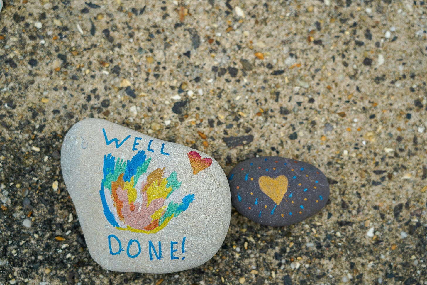 Photo of two painted stones on mottled beach sand. One says "well done" and the other has a heart