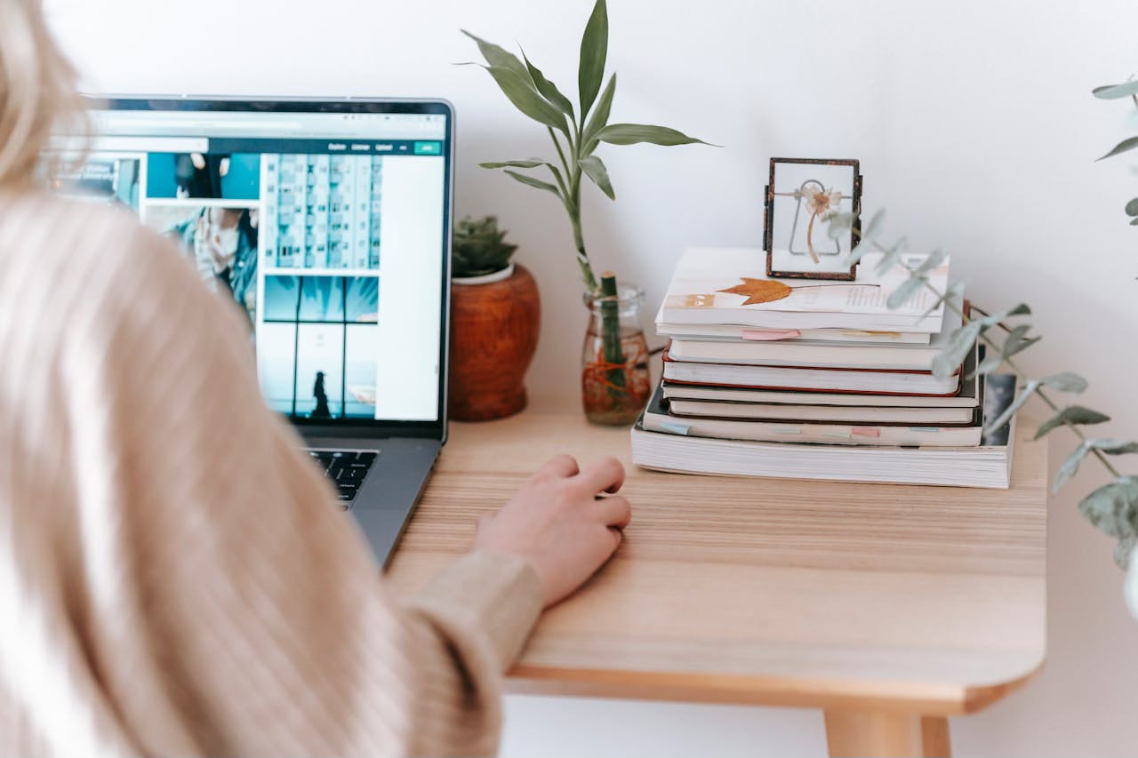Back view of a woman scrolling through images on a desktop computer with books and a plant sitting next to the computer on a light wood desk.