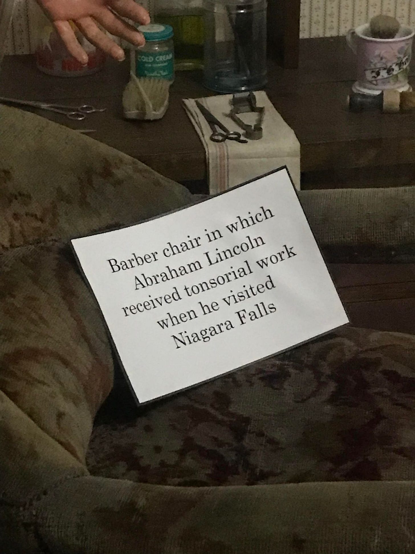 An old, grey-green velvet chair with a sign that reads 'barber chair in which Abraham Lincoln received tonsorial work when he visited Niagara Falls'