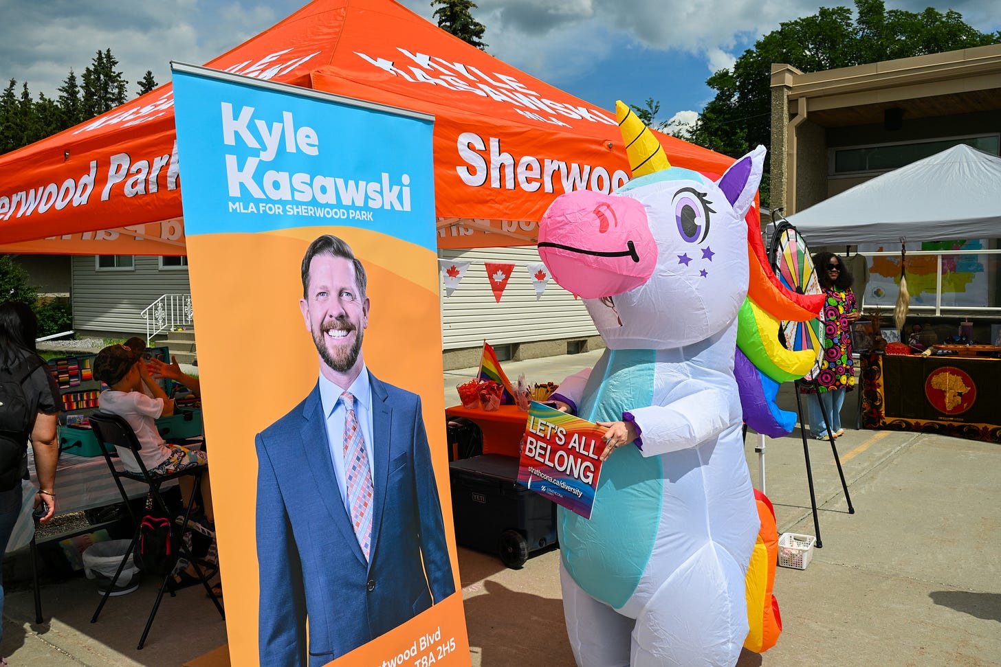 A banner with Kyle Kasawski's image on it stands in front of a bright orange event tent in front of the Strathcona county museum and a person in a rainbow unicorn costume holds a sign saying Let's all belong.