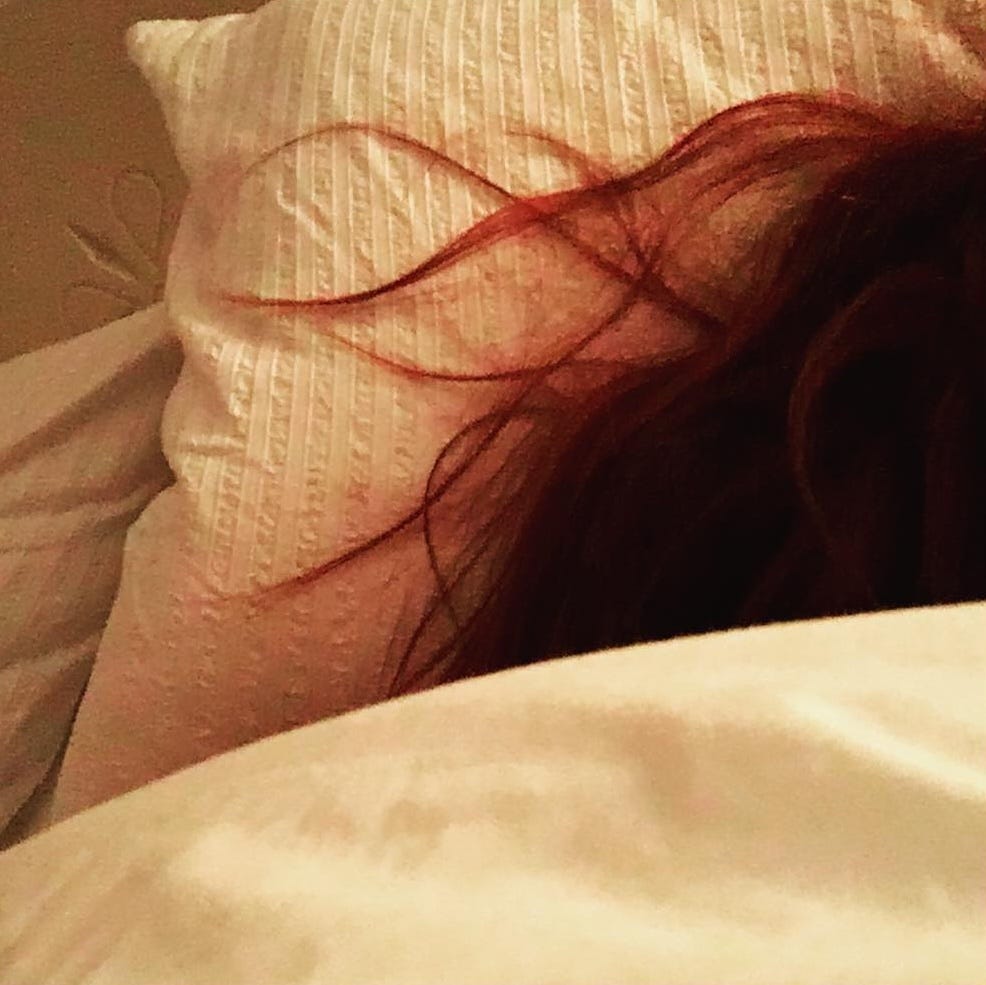 Red hair fans out across a white pillowcase, with a edge of a white duvet in the foreground.