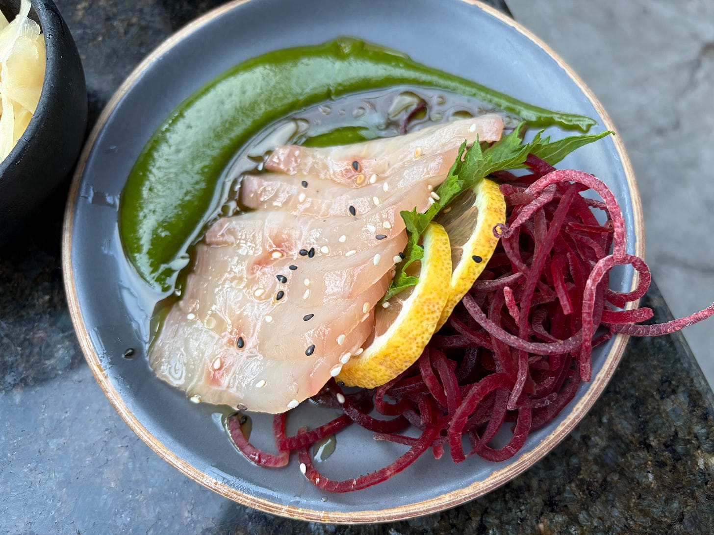 A top-down close-up shot of kanpachi sashimi presented on a dark grey ceramic dish. Thin, translucent slices of fish topped with black and white sesame seeds are nestled alongside a nest of spiralized red beets, and a slice of lemon, all resting in a pool of bright green arugula chimichurri sauce.