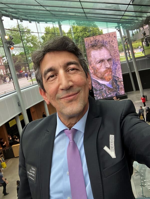 A middle-aged man with dark hair and eyes smiles at the camera in a museum and in front of a large video screen of Vincent van Gogh. A middle-aged man with dark hair and eyes smiles at the camera in a museum and in front of a large video screen of Vincent van Gogh.