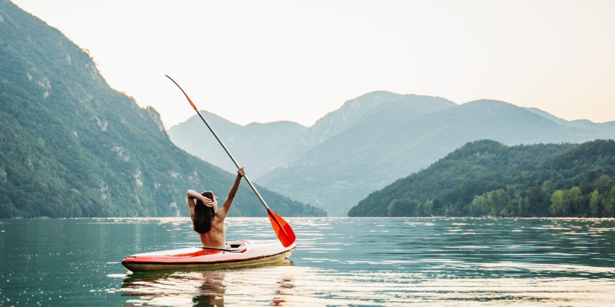A nude woman in a kayak seen from behind raises her oar, triumphantly taking in a nature scene.