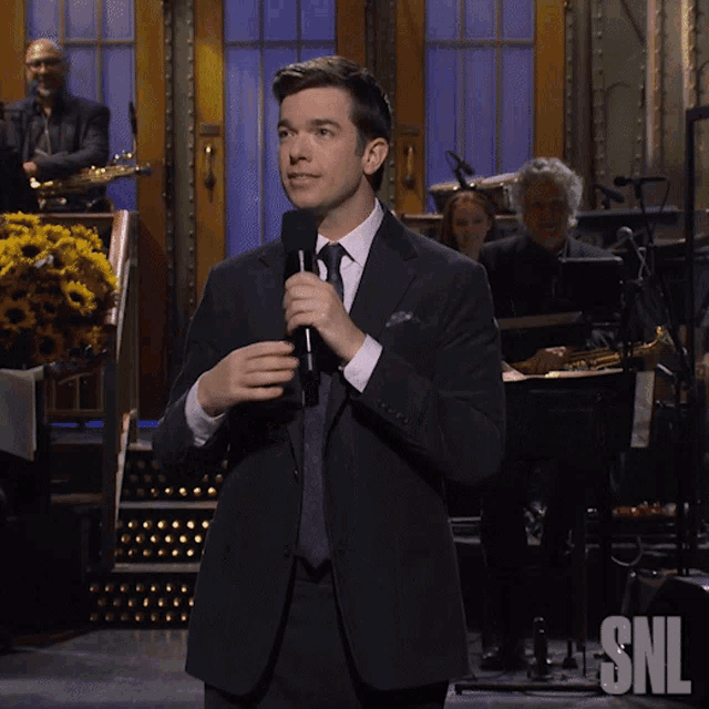 a man in a suit and tie holds a microphone in front of a snl sign a man in a suit and tie holds a microphone in front of a snl sign