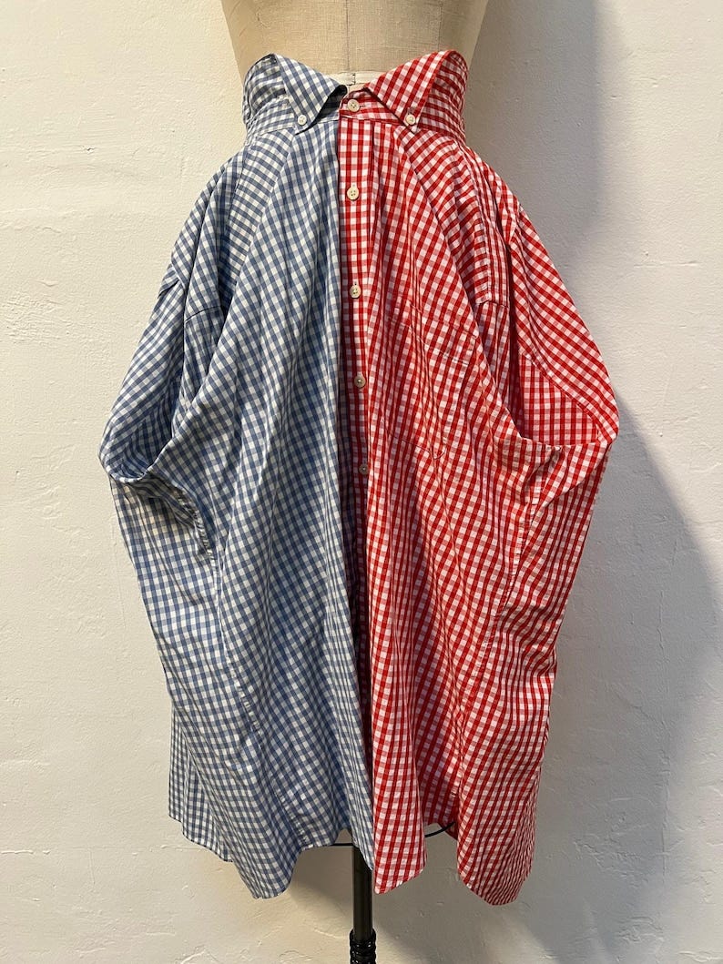 May include: A half-red and half-blue gingham midi skirt made from repurposed shirts. The skirt features a unique design with a center seam separating the two colors.  The fabric is a classic gingham check pattern, with a slightly A-line silhouette. The skirt's hemline falls to mid-calf length. The skirt is shown on a mannequin against a neutral background.